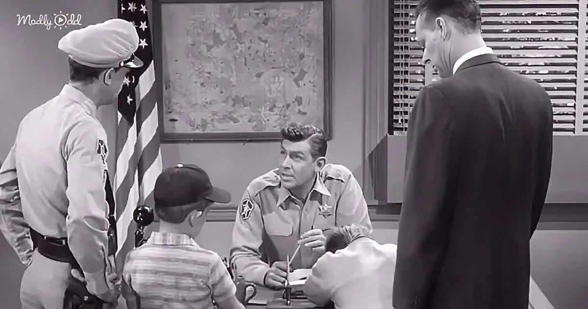A man in a sheriff's uniform sits at a desk, talking to a boy and another man in a suit, with a flag and map in the...