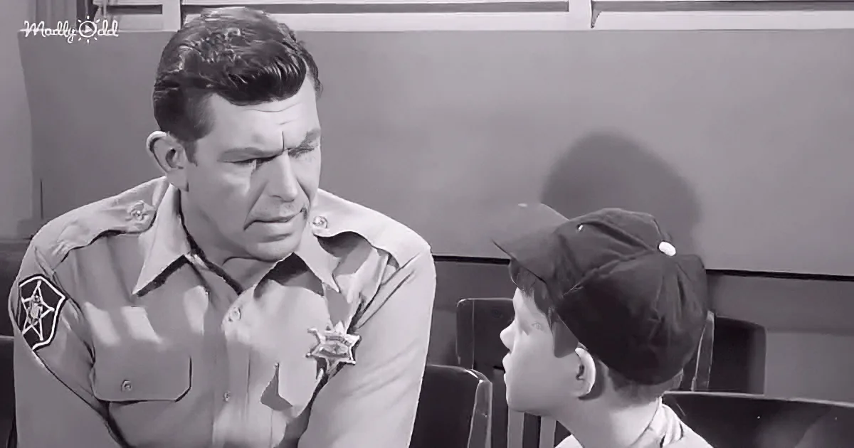 A sheriff in uniform speaks to a young boy in a baseball cap, both seated.