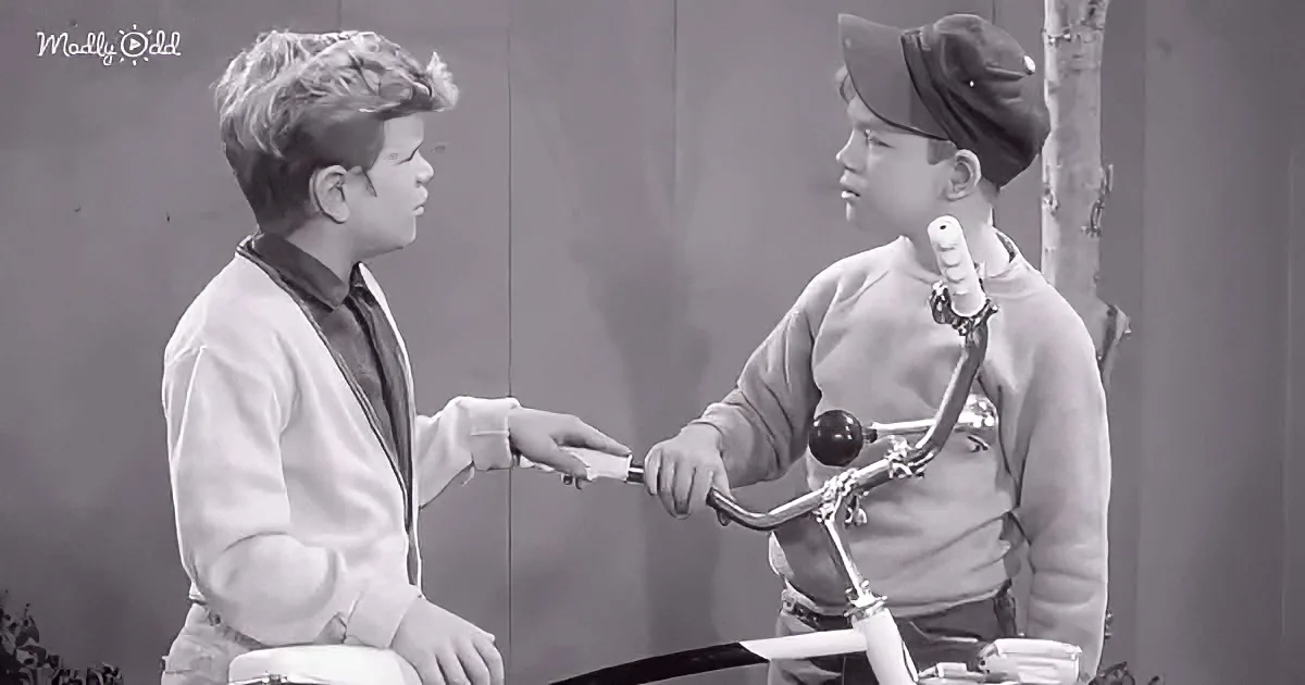 Two boys, one on a bike, exchange a small object, possibly a toy or a piece of candy.