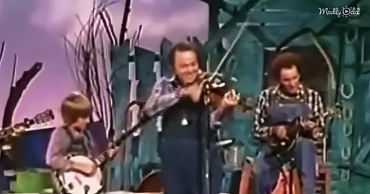 A man in a dark shirt plays violin on stage with two other musicians.