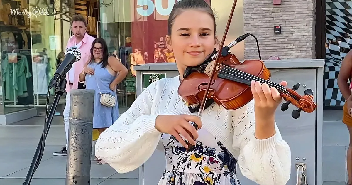A young girl in a white sweater and floral skirt plays the violin in front of a microphone, with people in the background.