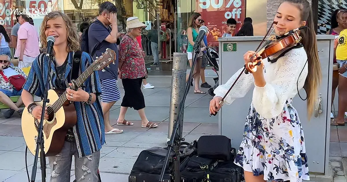 A young man plays guitar and sings while a woman plays violin in a bustling urban setting.
