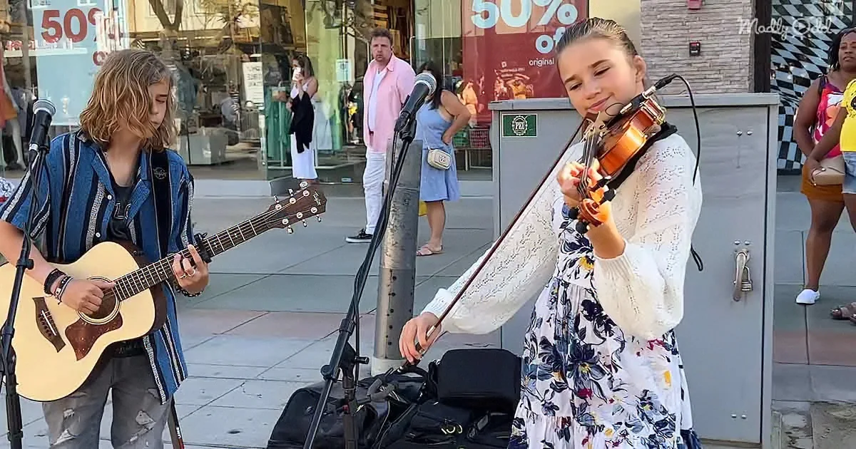 A young woman plays violin while a boy strums guitar on a city sidewalk, with passersby in the background.
