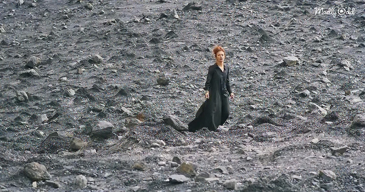 A woman in a black dress walks through a rocky, barren landscape.