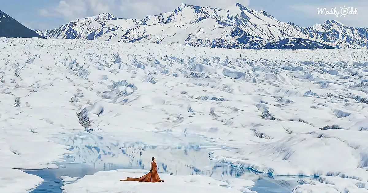 A woman in a flowing brown dress stands on a glacier, with snow-covered mountains in the background.