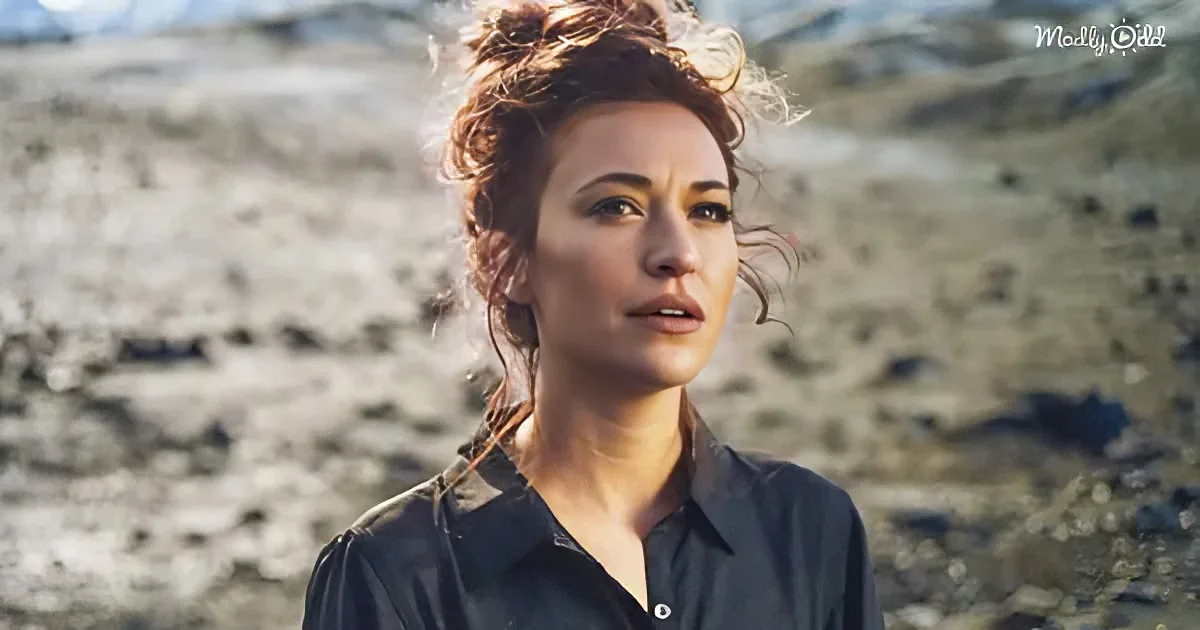 A woman with tousled hair and a dark shirt stands in a barren, rocky landscape.