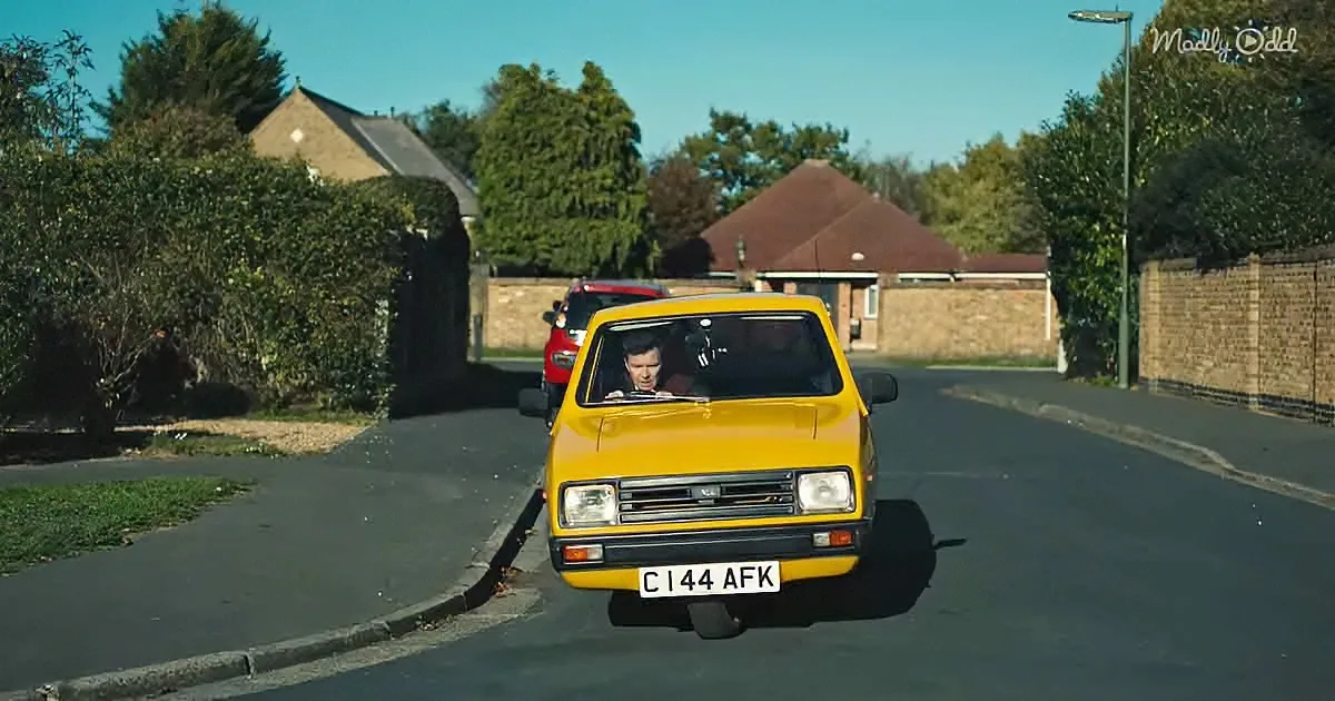 A yellow classic car drives down a suburban street with a person inside, surrounded by houses and greenery.
