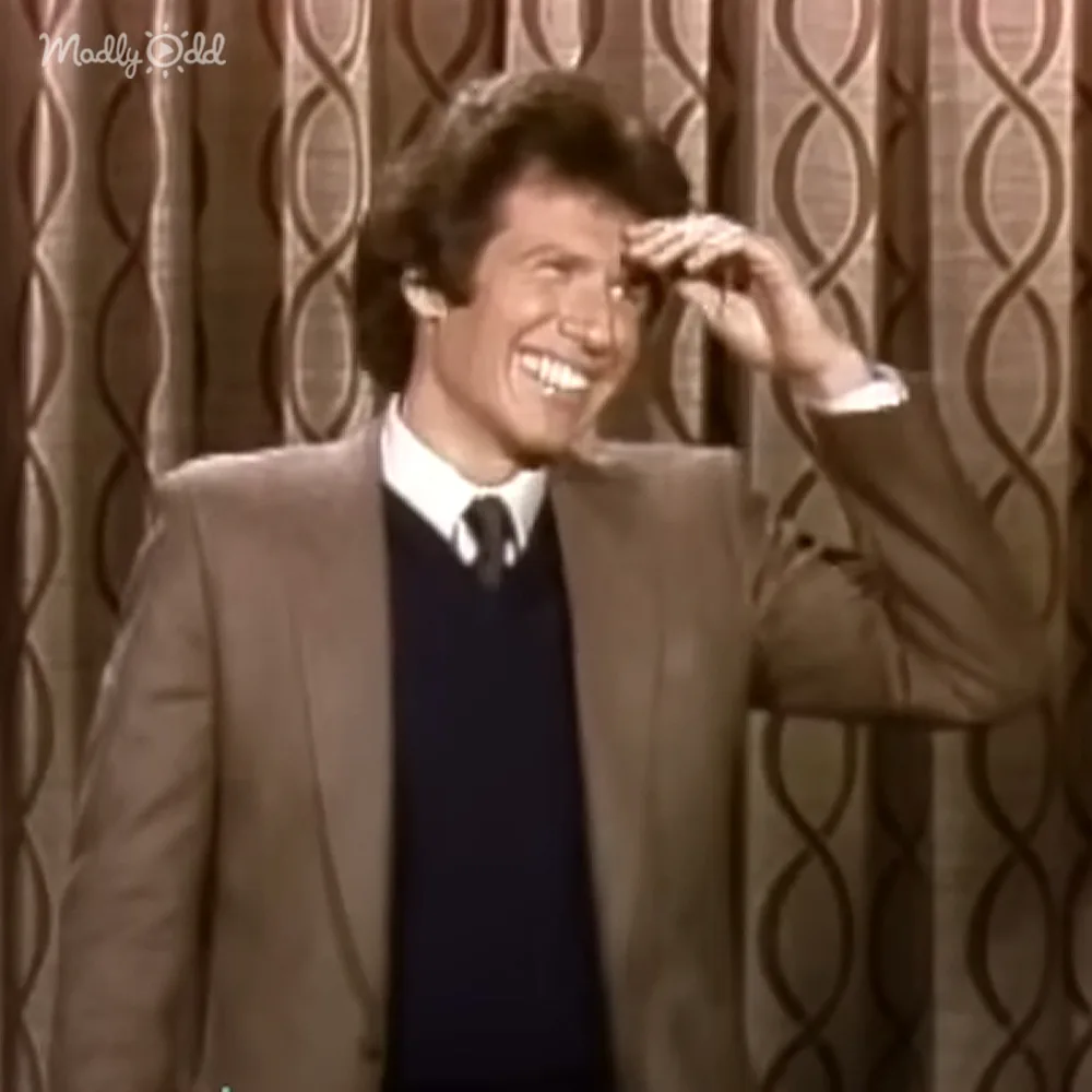 A man in a suit and tie smiles widely, holding his hand to his head, against a patterned curtain backdrop.