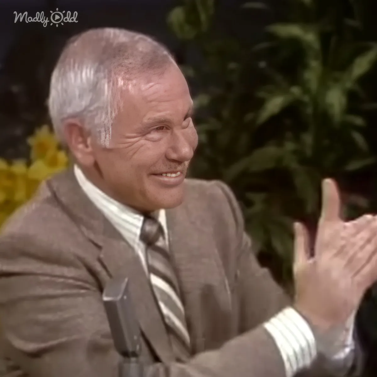 A man in a suit and tie is smiling and clapping, with a plant and yellow flowers in the background.