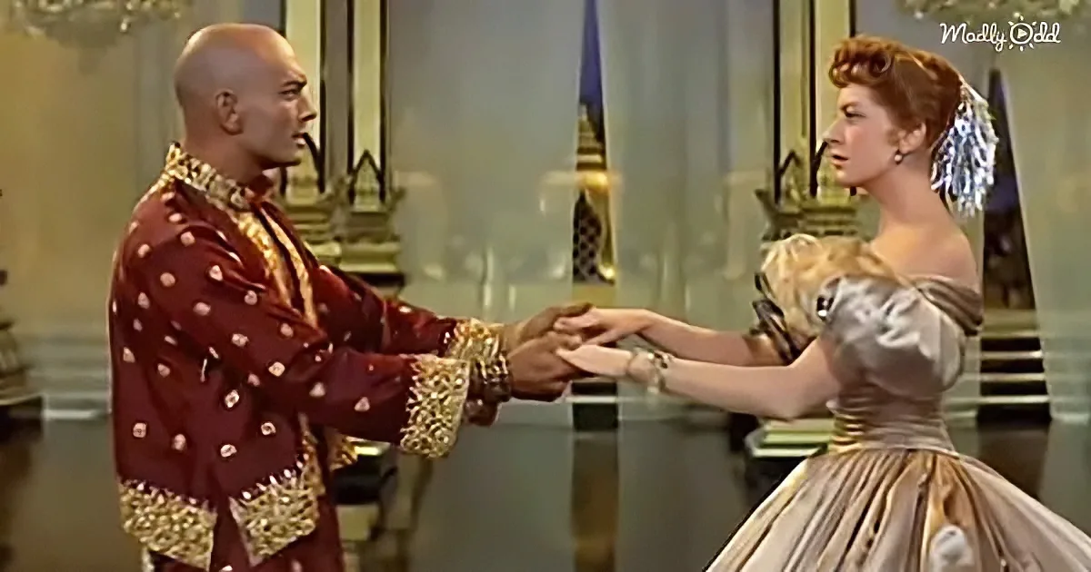 A man in a red, ornate robe and a woman in a silver dress hold hands in a grand, ornate room.