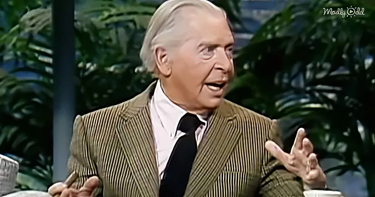 A man in a striped suit gestures while speaking, with tropical plants in the background.