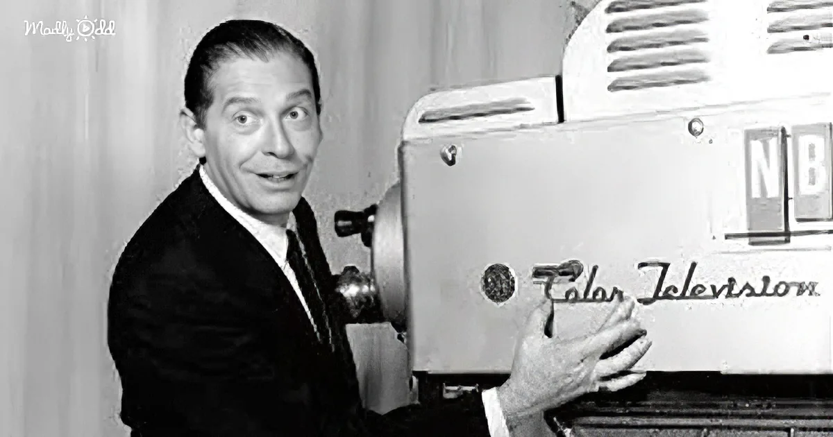 A man in a suit holds a vintage NBC color television camera, looking surprised.