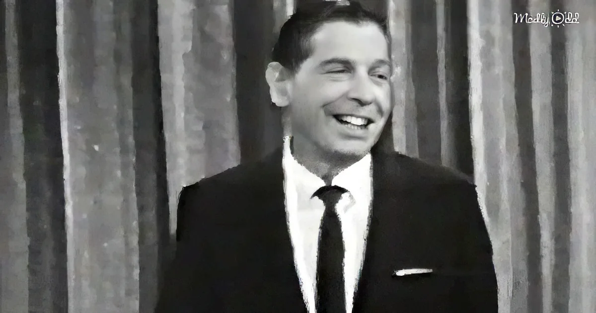 A man in a suit and tie smiles, standing in front of a curtain backdrop.