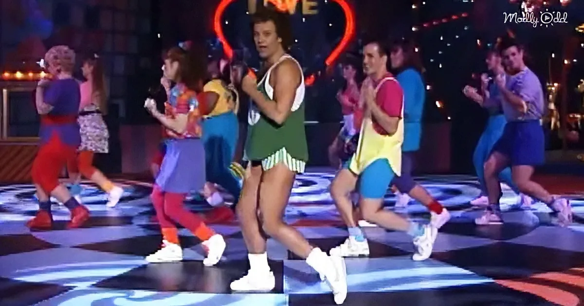 A group of people in colorful 80s-style outfits are dancing on a stage with a checkered floor.