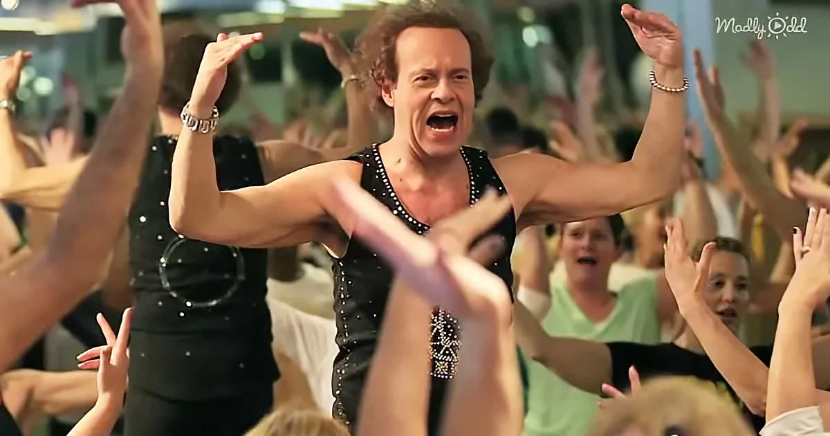 A man in a black sequined tank top leads a group in a synchronized dance with raised arms.