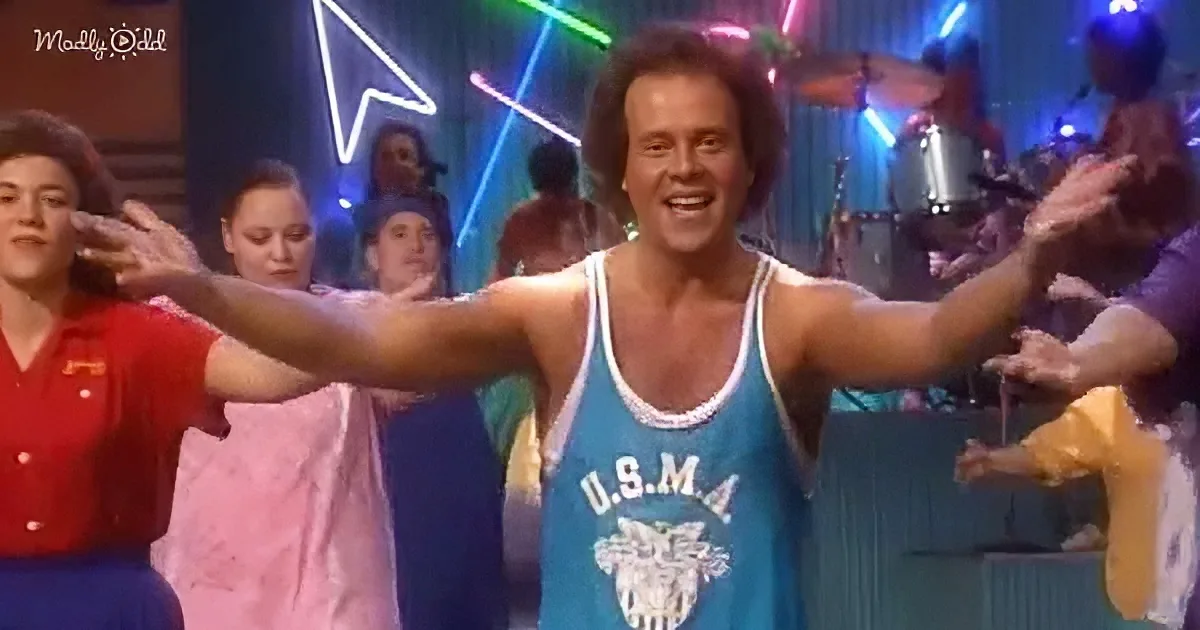 A man in a blue tank top with "U.S.M.A." on it is dancing with arms outstretched, surrounded by people and a band.