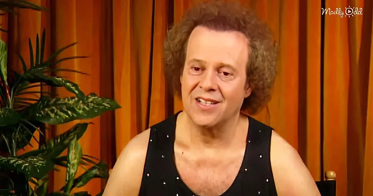 A person with curly hair and a black top with white dots is sitting in front of a wooden backdrop with a plant to the left.