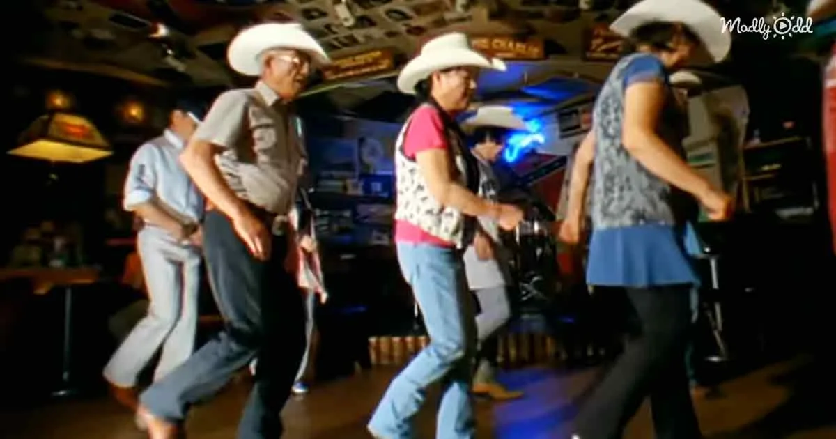 A group of people in cowboy hats and casual attire are dancing in a dimly lit bar with neon signs.