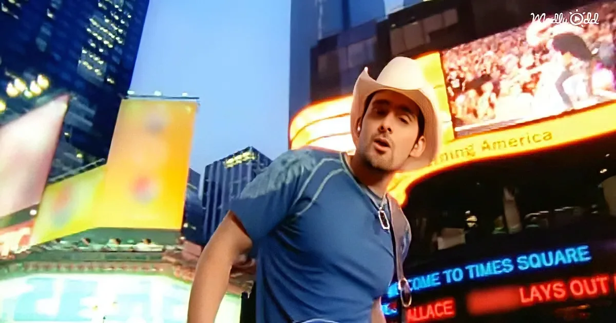 A man in a cowboy hat and blue shirt stands in front of a brightly lit Times Square backdrop.