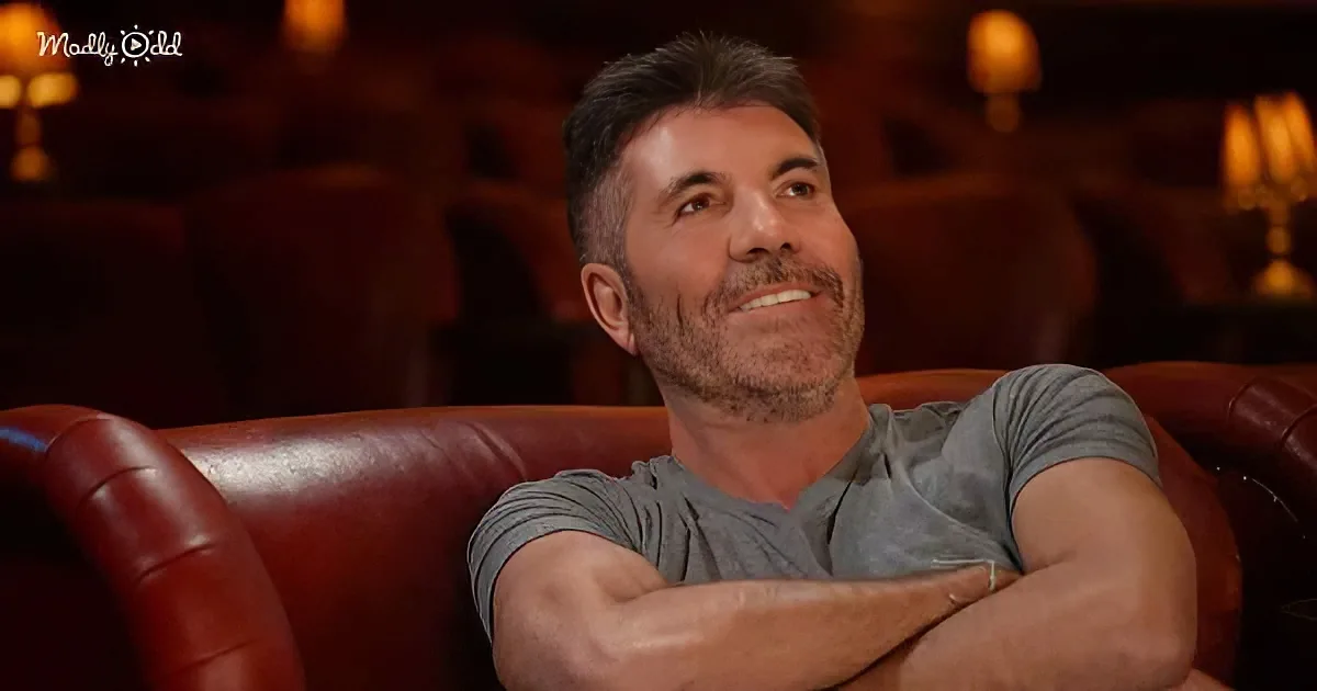 A man with a beard and gray shirt sits on a red leather couch, smiling and looking up.