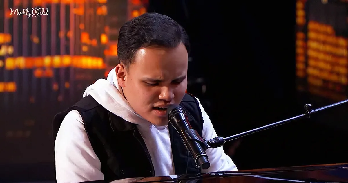 A person in a white hoodie and black vest sings into a microphone, with a piano and warm background lights.