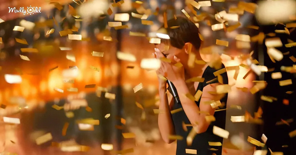 A woman in a black dress stands amidst golden confetti, holding her hands to her face.