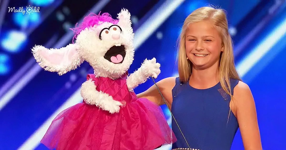 A young girl in a blue dress stands next to a puppet in a pink dress on a stage.