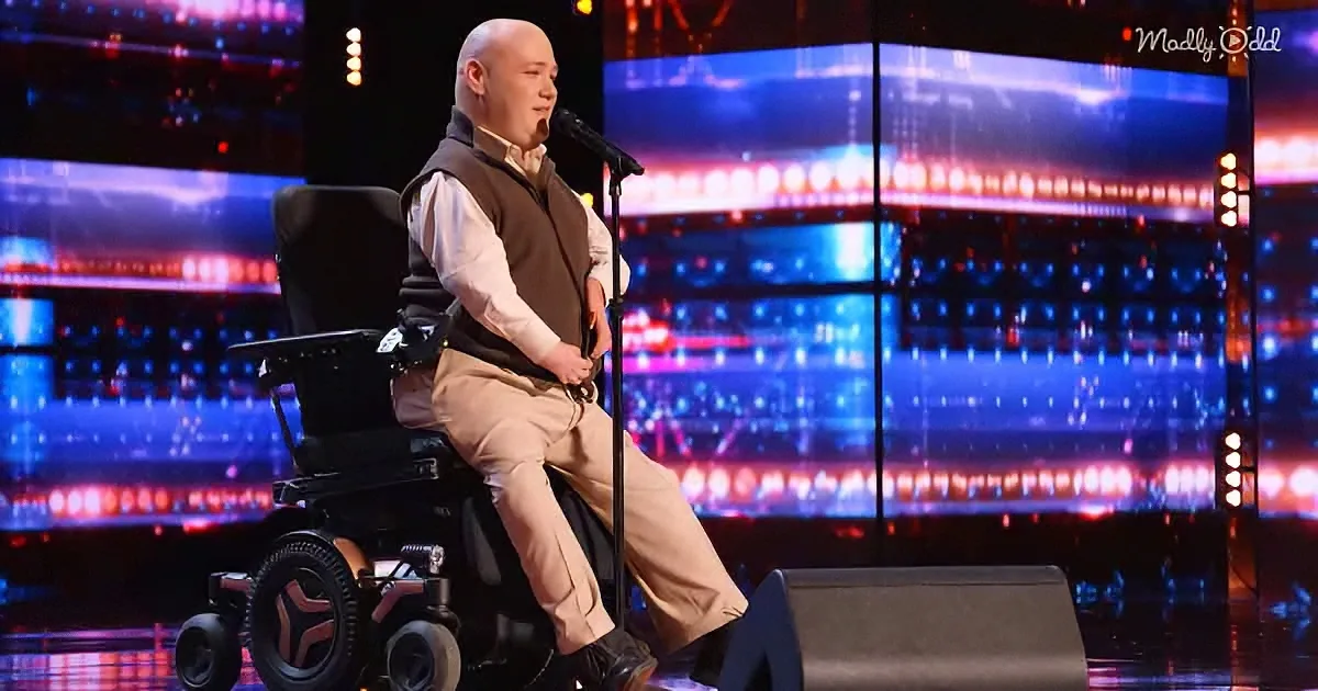 A man in a wheelchair stands on stage, holding a microphone, dressed in a vest and pants.