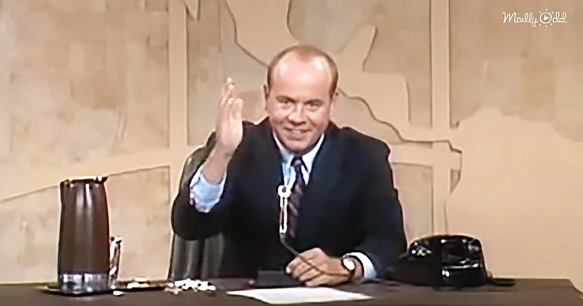 A man in a suit sits at a desk, gesturing with his right hand while holding a pen in his left.