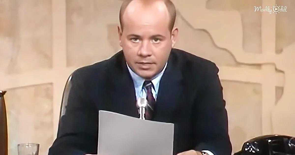 A man in a suit sits at a desk, holding a paper, with a beige background and a "Madly Odd" watermark.
