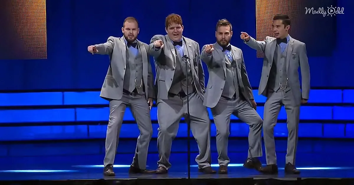 Four men in gray suits perform on stage, pointing forward with blue-lit background.