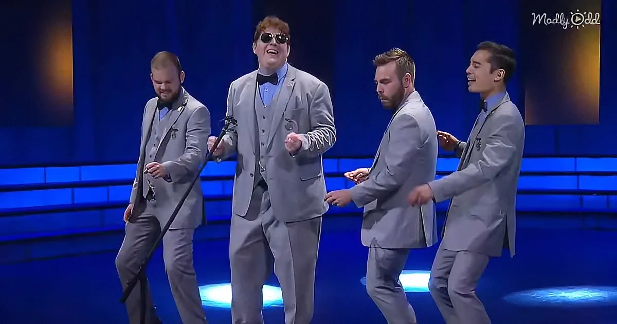 A man in a gray suit and sunglasses is dancing on stage with three other men in matching suits.
