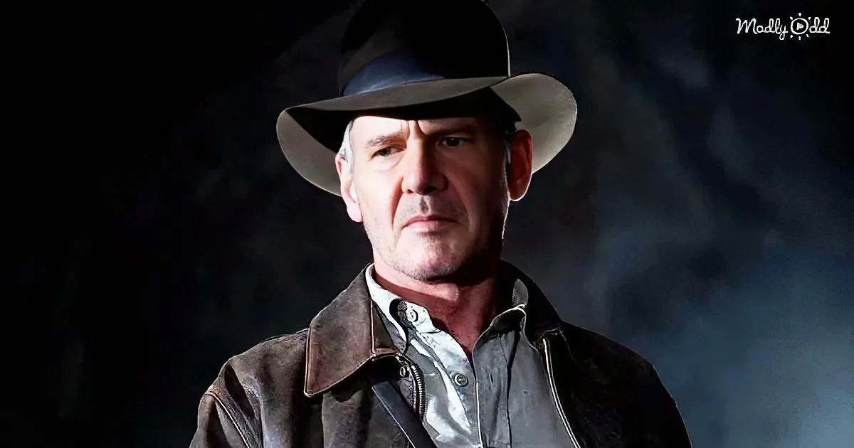 A man in a brown leather jacket and wide-brimmed hat stands against a dark background, looking serious.