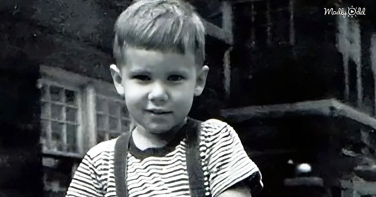 A young boy with short hair, wearing a striped shirt and suspenders, stands in front of a rustic house.