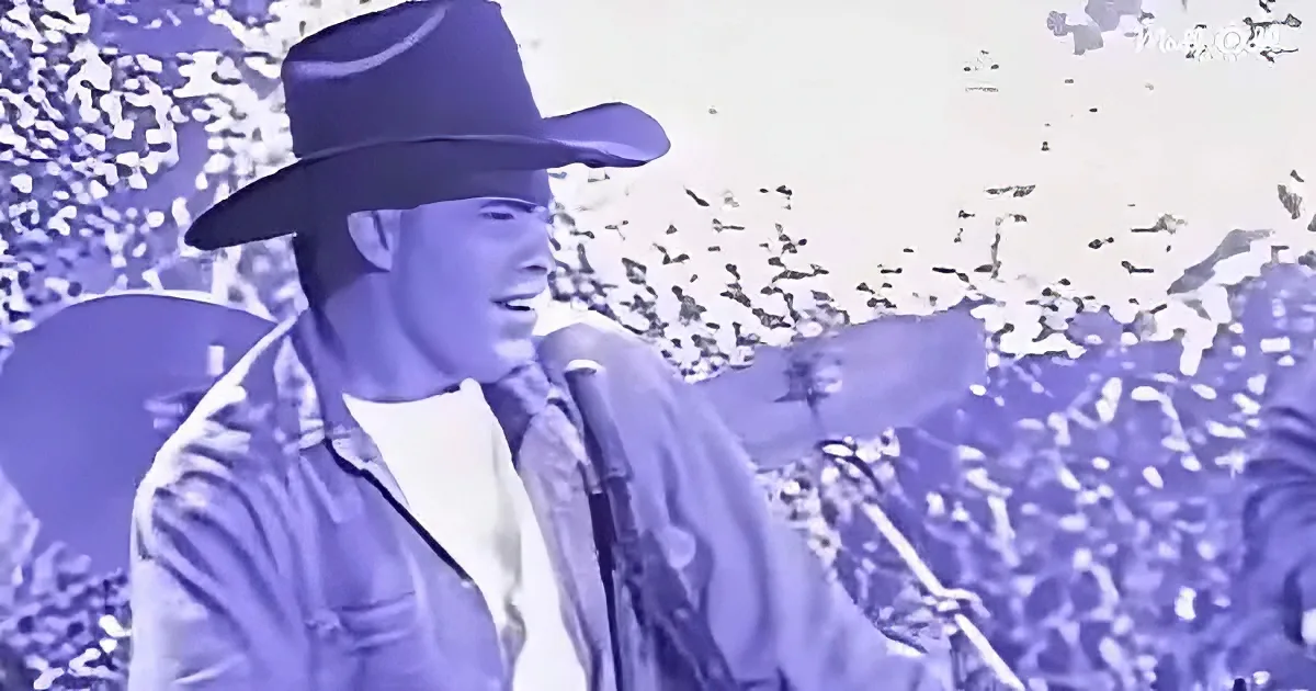 A man in a cowboy hat sings into a microphone, surrounded by a crowd.