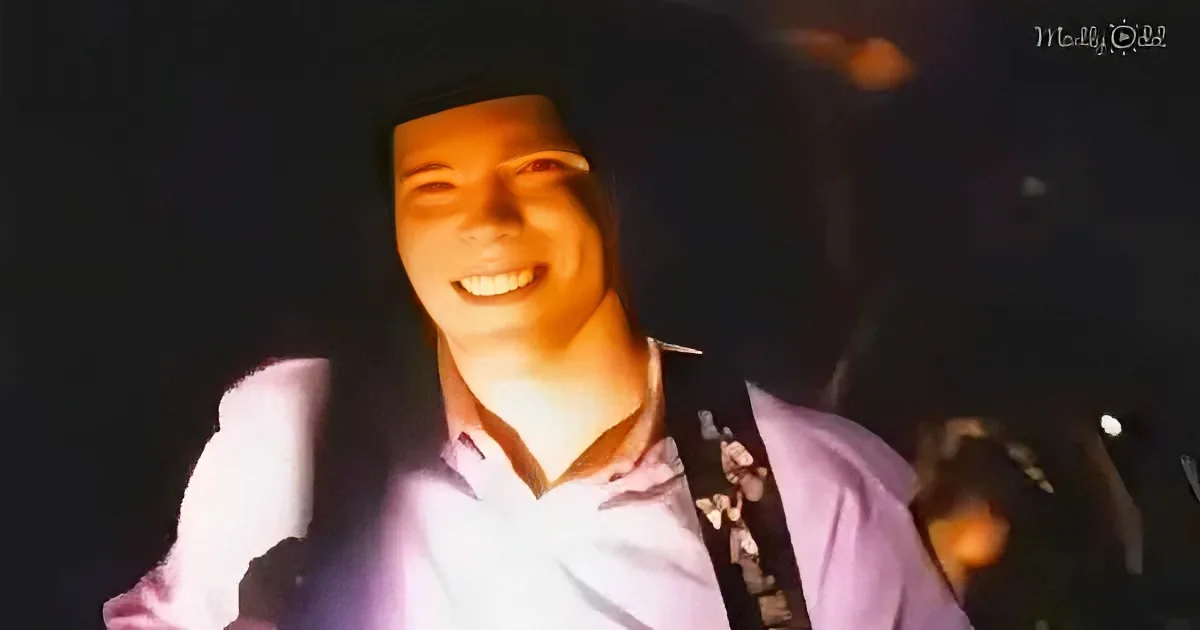 A smiling person in a pink shirt with a guitar strap, illuminated by stage lights.