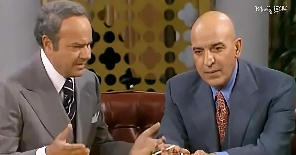 A man in a suit gestures while seated next to another man in a suit, both in a studio setting.
