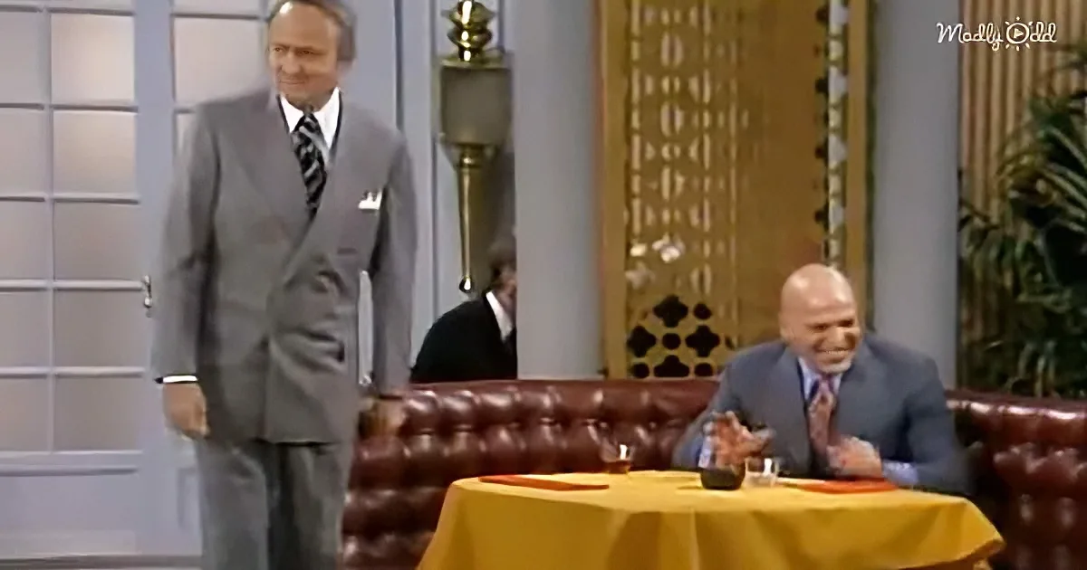 A man in a suit stands beside a seated man in a suit, both in a room with a yellow tablecloth and leather couch.