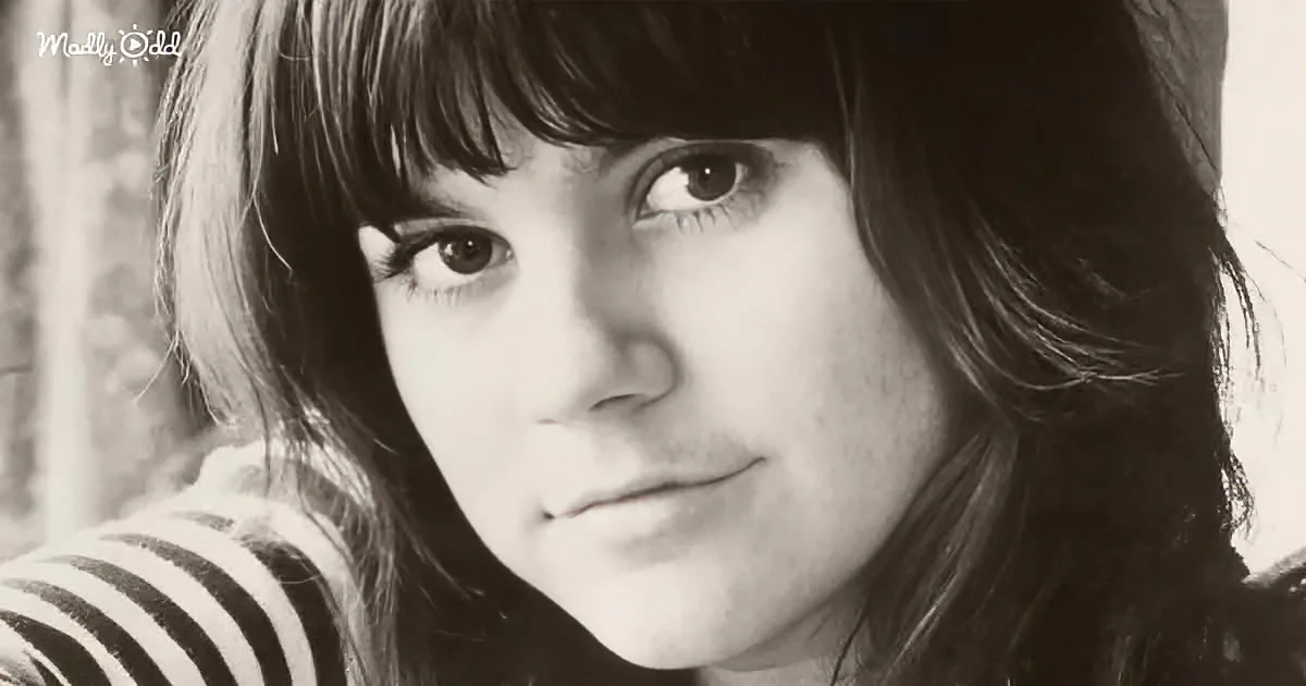 A person with long hair and bangs, wearing a striped shirt, looks directly at the camera in a black-and-white photo.