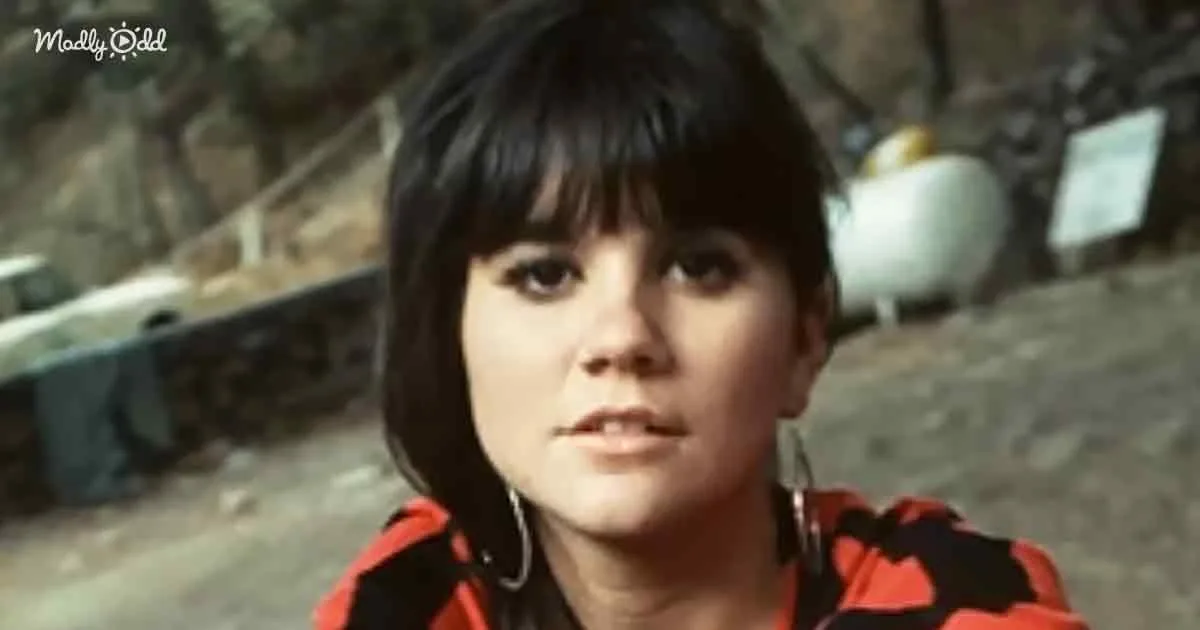A person with short dark hair and bangs, wearing a red and black outfit, stands outdoors with a rocky background.