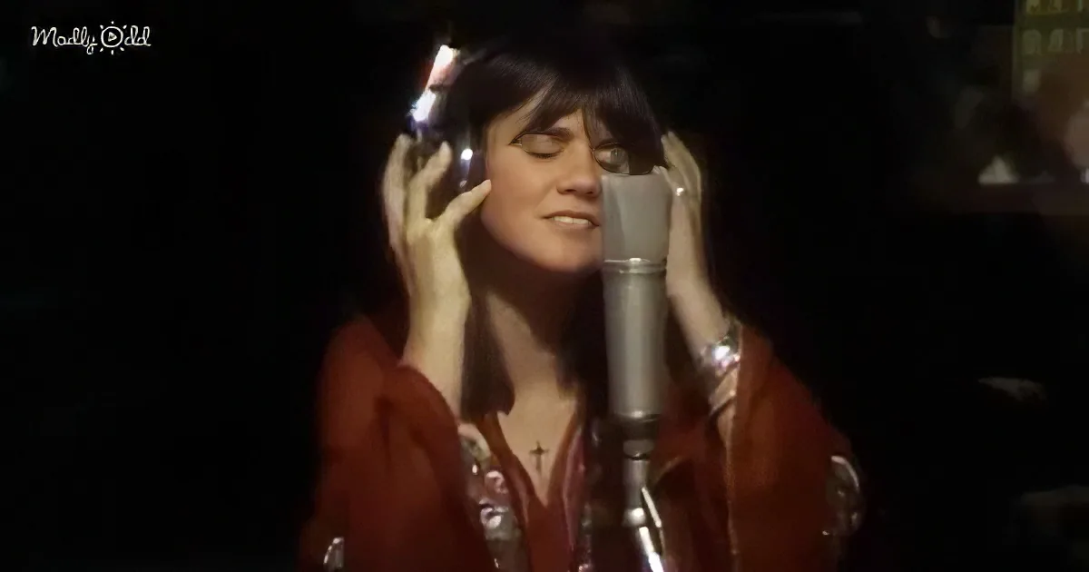 A woman sings into a microphone, holding headphones, wearing a red jacket and a cross necklace.