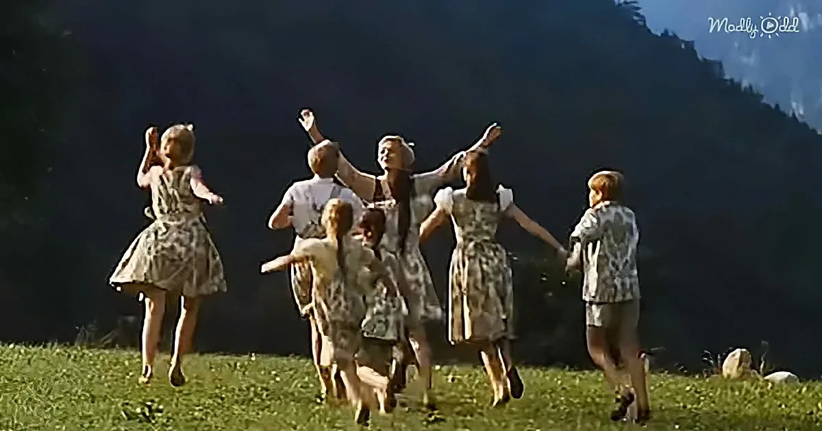 Children in vintage dresses and shirts dance joyfully on a grassy hillside against a dark mountain backdrop.