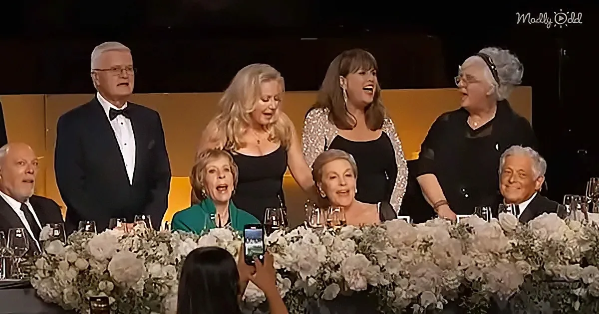 A group of people, including a woman in a black dress, are gathered around a table adorned with flowers and wine glasses.