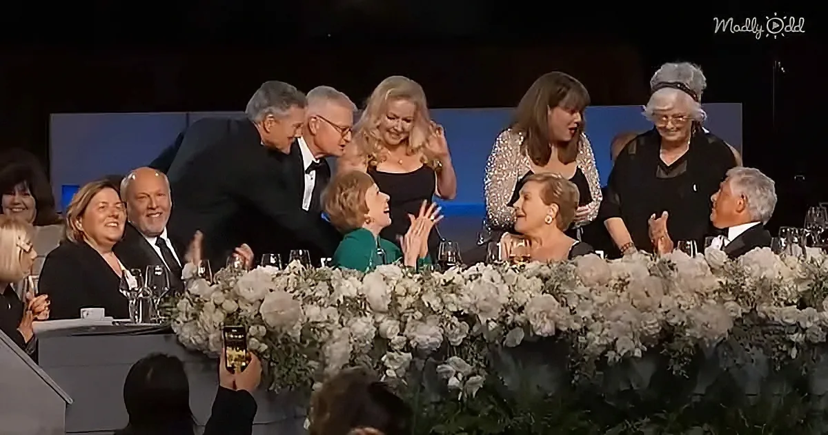 A man in a tuxedo leans over a table filled with flowers, engaging with a group of people at what appears to be a formal...