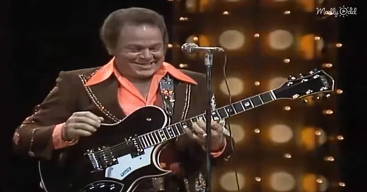 A man in a brown suit and orange shirt smiles while playing a black electric guitar on stage.