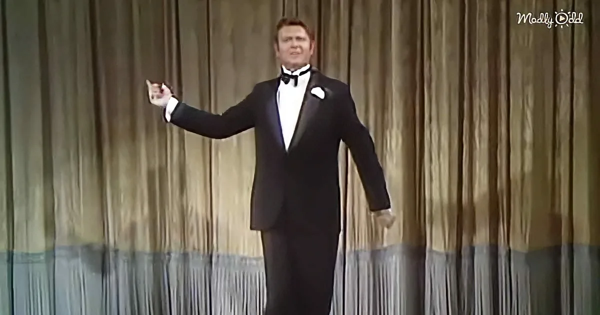 A man in a tuxedo stands on stage, gesturing with his right hand.