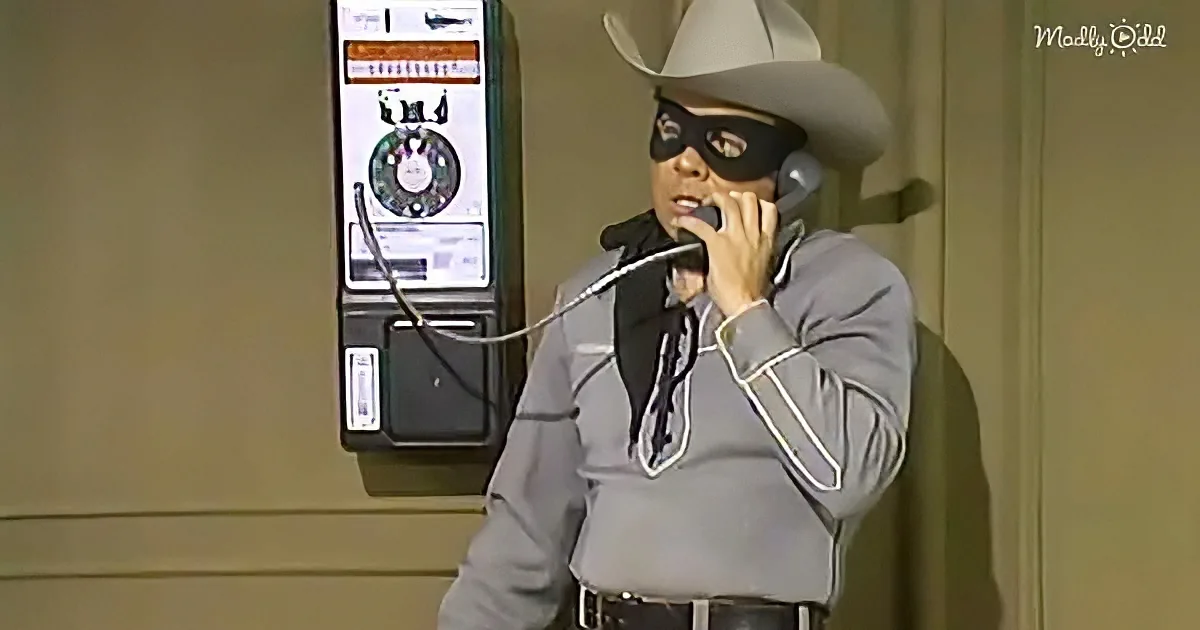 A person in a cowboy outfit and mask talks into a microphone, with a payphone in the background.