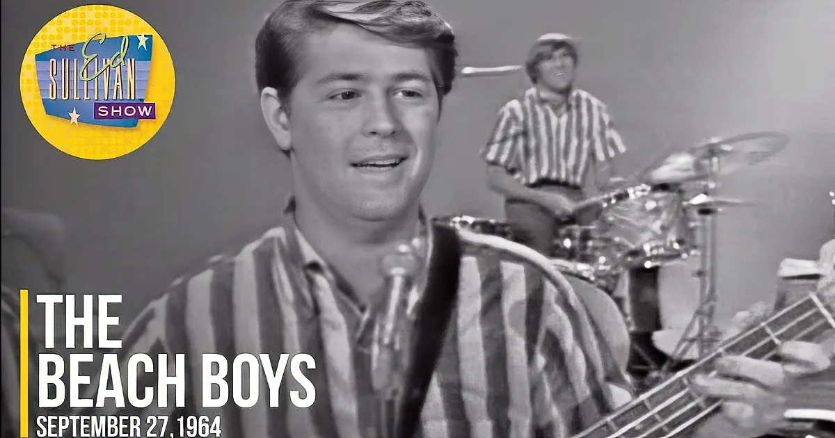 A young man in a striped shirt plays a bass guitar, smiling, with a drummer in the background.
