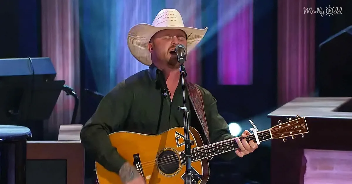 A man in a cowboy hat sings into a microphone while playing an acoustic guitar on stage.