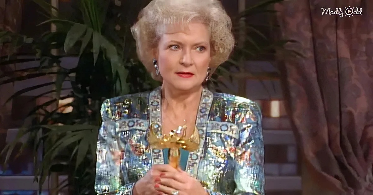 An older woman with blonde hair, wearing a colorful patterned jacket, holds a trophy in a studio setting with plants in...